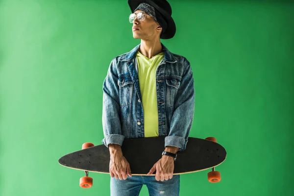 Stylish mixed race man in denim posing with skateboard on green screen — Stock Photo