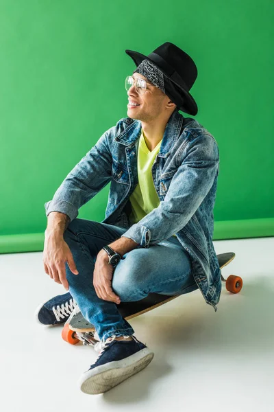 Стильный смешанный гонщик в джинсах сидит на скейтборде и улыбается на зеленом — стоковое фото