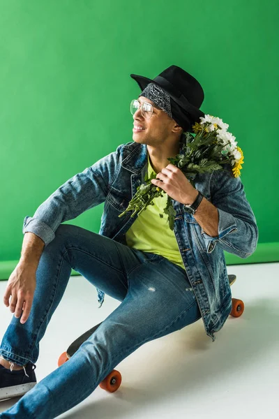 Homem de raça mista elegante em ganga sentado no skate com flores e posando no verde — Fotografia de Stock