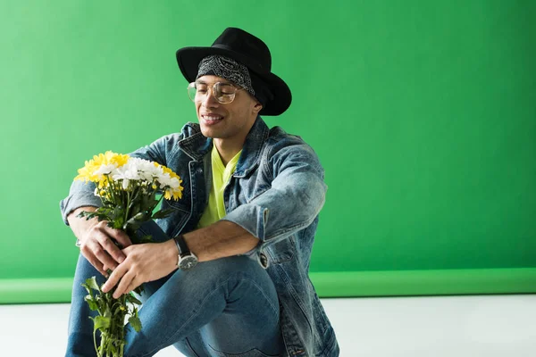 Stylish mixed race man in glasses sitting, holding flowers and smiling on green — Stock Photo