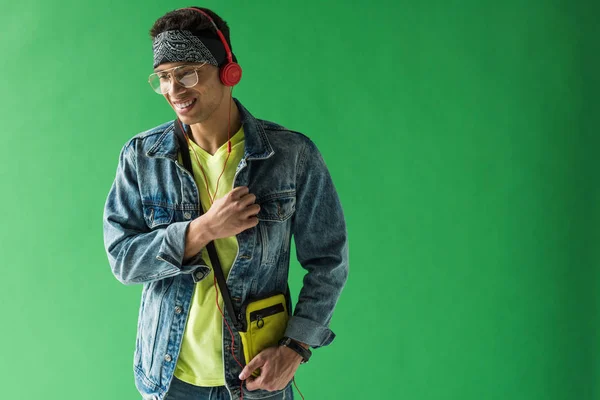 Apuesto hombre de raza mixta en auriculares escuchando música y sonriendo en la pantalla verde con espacio de copia - foto de stock