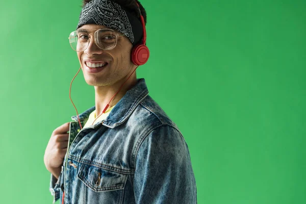 Apuesto hombre de raza mixta en los auriculares mirando a la cámara y sonriendo mientras escucha música en la pantalla verde - foto de stock