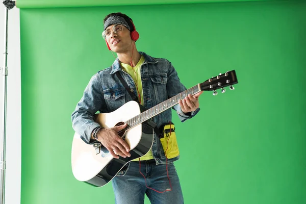 Handsome mixed race man in denim and headphones playing acoustic guitar on green — Stock Photo