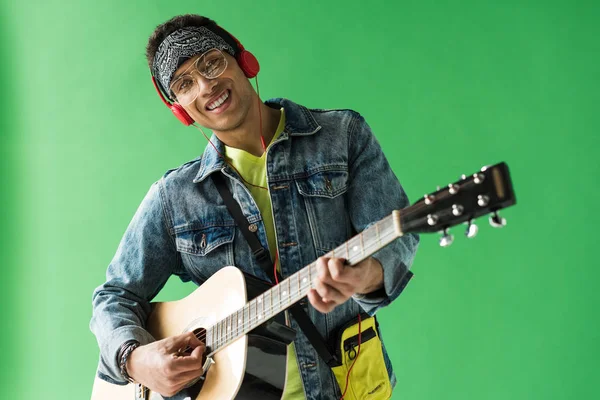 Bel homme métis en denim et écouteurs regardant la caméra tout en jouant de la guitare acoustique sur écran vert — Photo de stock