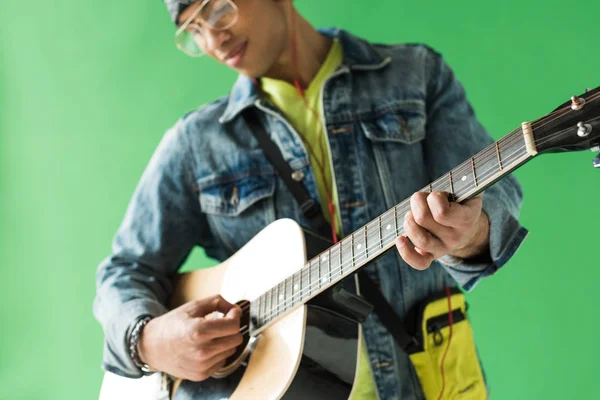 Селективное внимание человека смешанной расы в джинсах, играющего на акустической гитаре на зеленом экране — стоковое фото