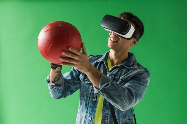 Hombre de raza mixta en auriculares de realidad virtual sosteniendo el baloncesto y sonriendo en la pantalla verde - foto de stock