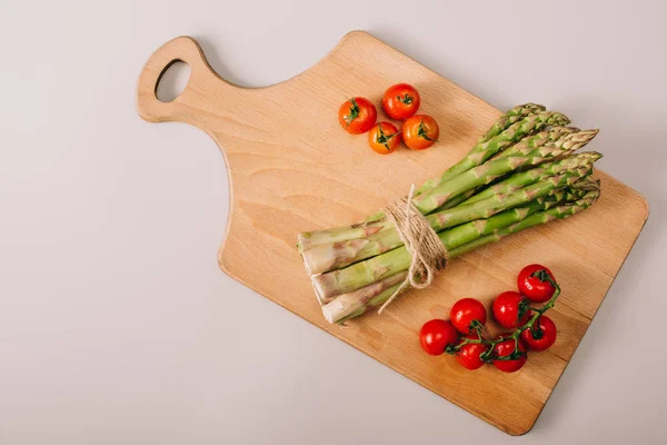 Draufsicht auf grünen, ungekochten Spargel und Kirschtomaten auf Holzschneidebrett auf grauem Hintergrund — Stockfoto