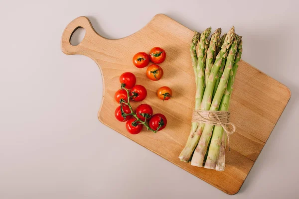 Vista superior de espargos verdes crus e tomates cereja em tábua de corte de madeira em fundo cinza — Fotografia de Stock