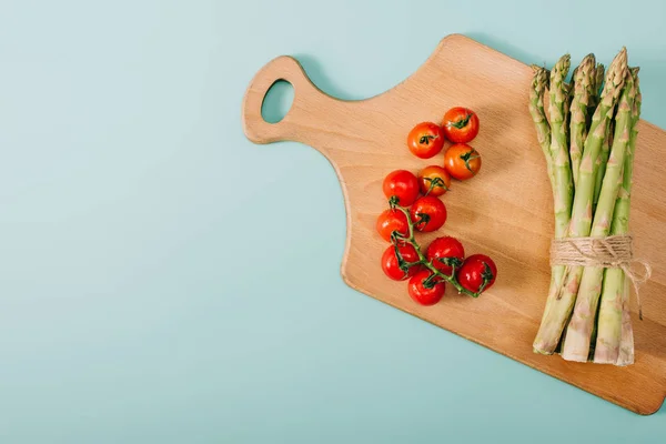 Top view of green raw asparagus and cherry tomatoes on wooden cutting board on blue background — Stock Photo