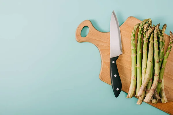 Vista dall'alto di asparagi grezzi verdi su tagliere in legno con coltello su fondo blu — Foto stock