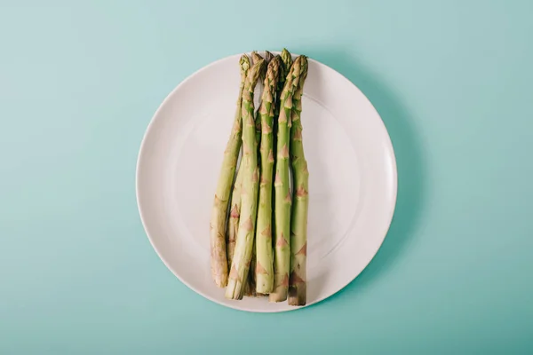 Vue de dessus des asperges crues vertes sur plaque blanche sur fond bleu — Photo de stock