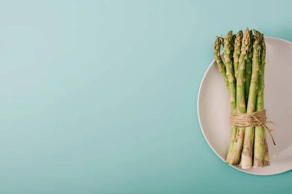 Vue de dessus des asperges crues vertes attachées à la corde sur plaque blanche sur fond bleu avec espace de copie — Photo de stock