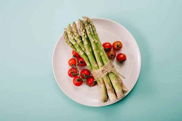 Top view of green raw asparagus and cherry tomatoes on white plate on blue background — Stock Photo