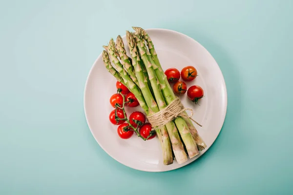 Draufsicht auf grünen rohen Spargel und Kirschtomaten auf weißem Teller auf blauem Hintergrund — Stockfoto