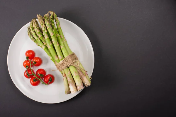 Draufsicht auf grünen Spargel und Kirschtomaten auf Teller auf schwarzem Hintergrund — Stockfoto