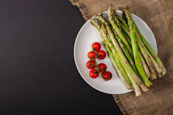 Draufsicht auf grünen Spargel und Kirschtomaten auf Teller in der Nähe von Sacktuch auf schwarzem Hintergrund — Stockfoto