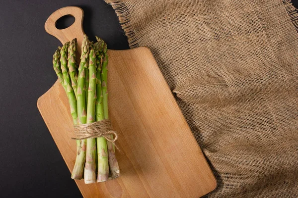 Vista dall'alto di mazzo di asparagi verdi legati con corda su tagliere di legno vicino al sacco — Foto stock