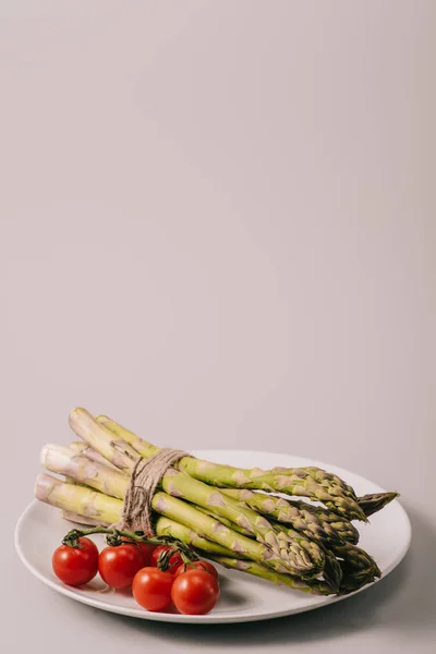 Кучка зеленой сырой спаржи, привязанной веревкой и помидорами черри на белой тарелке на сером фоне — стоковое фото