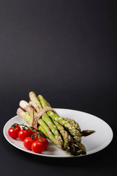 Bunch of green uncooked asparagus tied with rope and cherry tomatoes on white plate on black background — Stock Photo