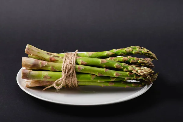 Manojo de espárragos verdes sin cocer atados con cuerda en plato blanco sobre fondo negro - foto de stock