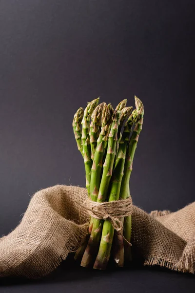 Mazzo di asparagi verdi non cotti legati con corda vicino al sacco su sfondo nero — Foto stock