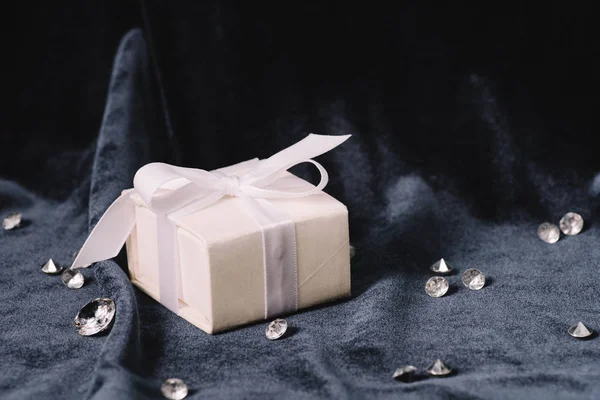 Gift box with white ribbon and bow near pure diamonds on blue cloth — Stock Photo
