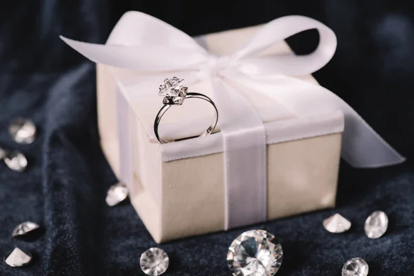 Selective focus of engagement ring on gift box with bow near shiny diamonds on blue cloth — Stock Photo