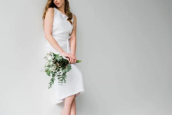 Vista ritagliata della donna in vestito che tiene mazzo di fiori con foglie di eucalipto su bianco — Foto stock