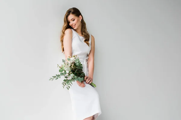 Mulher sorridente em vestido segurando buquê de flores com folhas de eucalipto no branco — Fotografia de Stock