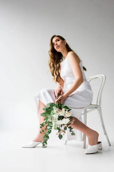 Low angle view of woman in dress sitting on chair and holding bouquet of flowers with eucalyptus leaves on white — Stock Photo