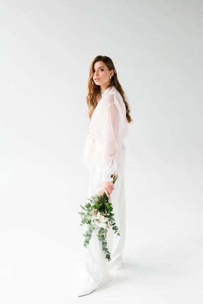 Atractiva mujer sosteniendo flores con hojas de eucalipto verde mientras está de pie sobre blanco - foto de stock