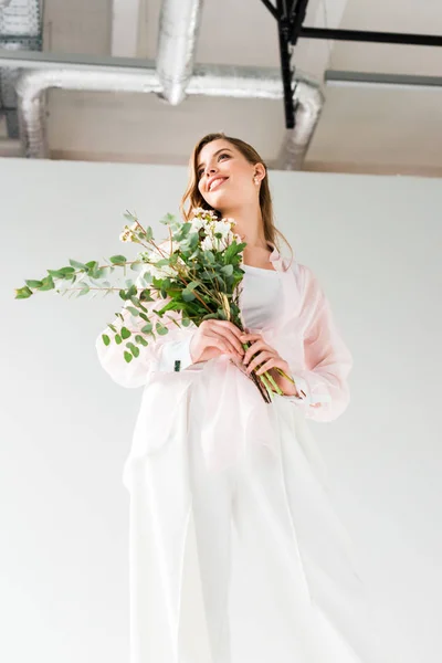 Low angle view of happy woman holding flowers with green eucalyptus leaves while standing on white — Stock Photo