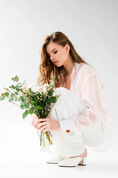 Attraktive Frau, die Blumen mit grünen Eukalyptusblättern betrachtet, während sie auf weiß sitzt — Stockfoto