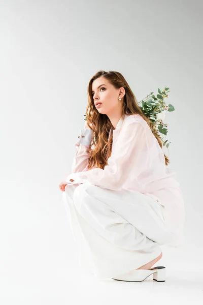 Atractiva mujer sosteniendo flores con hojas de eucalipto verde detrás de la espalda mientras está sentado en blanco - foto de stock