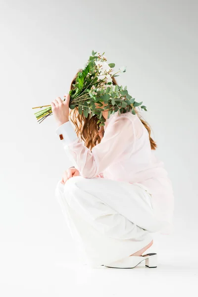 Chica cubriendo la cara con flores con hojas de eucalipto verde mientras está sentado en blanco - foto de stock