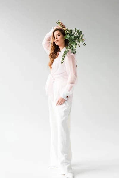 Hermosa mujer joven sosteniendo flores con hojas de eucalipto verde detrás de la espalda mientras está de pie en blanco - foto de stock