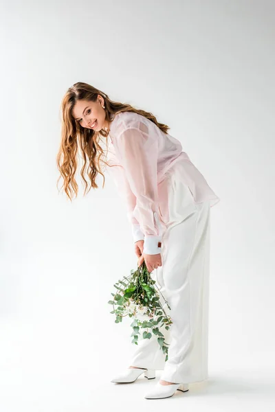 Happy and stylish young woman holding flowers with eucalyptus leaves on white — Stock Photo