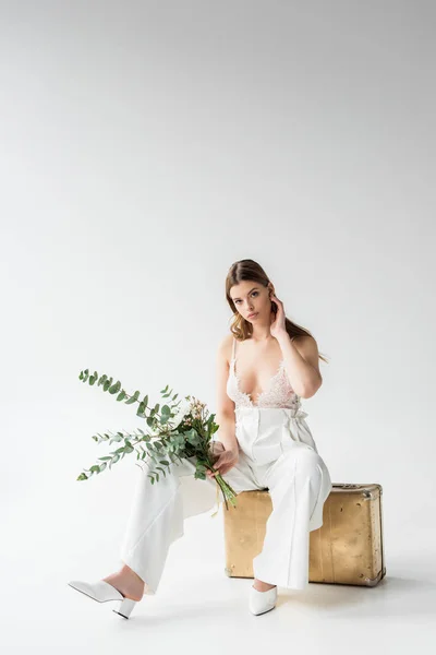 Стильна молода жінка сидить на дорожній сумці і тримає букет з квітами та листям евкаліпта на білому — стокове фото