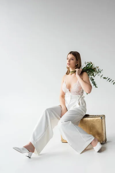 Jeune femme assise sur un sac de voyage et tenant un bouquet avec des fleurs et des feuilles d'eucalyptus sur blanc — Photo de stock