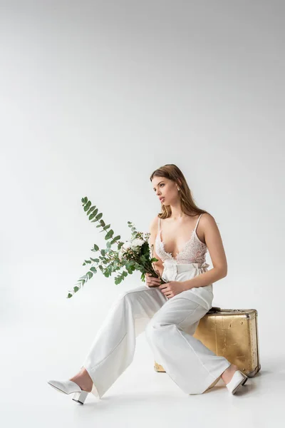 Woman sitting on travel bag and holding bouquet with flowers and eucalyptus leaves on white — Stock Photo