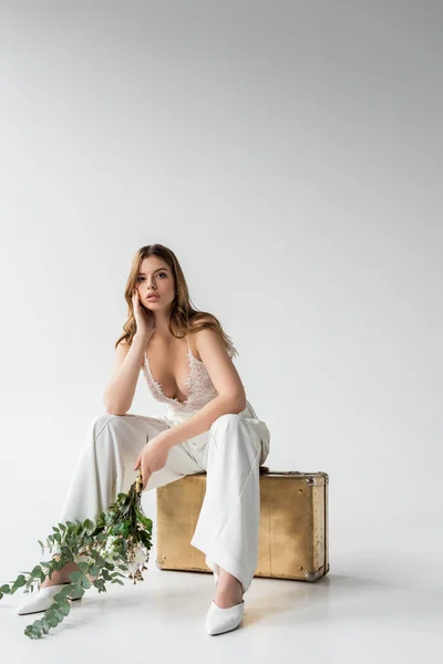 Girl sitting on travel bag and holding bouquet with flowers and eucalyptus leaves on white — Stock Photo