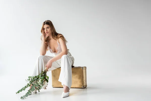 Задумчивая девушка сидит на дорожной сумке и держит букет с цветами и эвкалиптовыми листьями на белом — стоковое фото