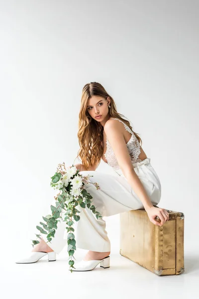 Jolie fille assise sur le sac de voyage et tenant bouquet avec des fleurs et des feuilles d'eucalyptus sur blanc — Photo de stock