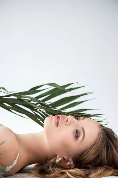 Bella giovane donna sdraiata vicino a foglie di palma tropicale su bianco — Foto stock