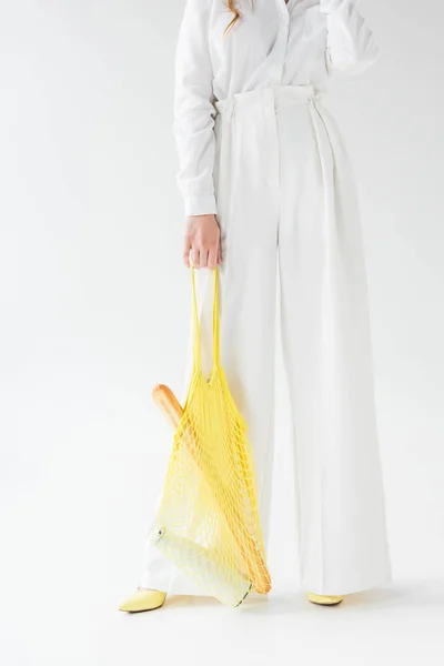 Vista recortada de la mujer con baguette y botella de leche en bolsa de hilo amarillo de pie sobre blanco - foto de stock