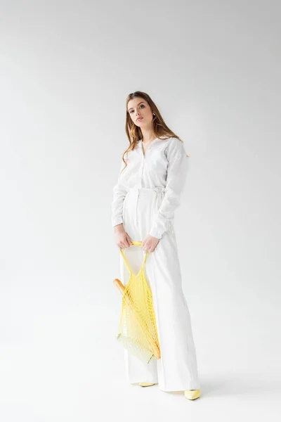 Jovem bonita segurando saco de corda amarela com baguete e garrafa de leite no branco — Fotografia de Stock
