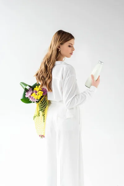 Attractive woman holding bottle of milk and yellow string bag with wildflowers on white — Stock Photo