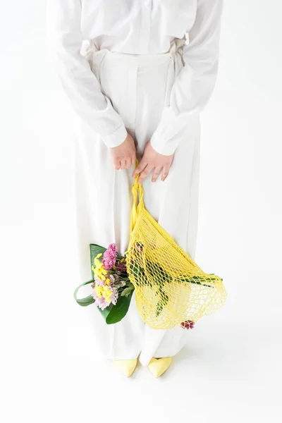 Cropped view of woman holding yellow string bag with wildflowers while standing on white — Stock Photo