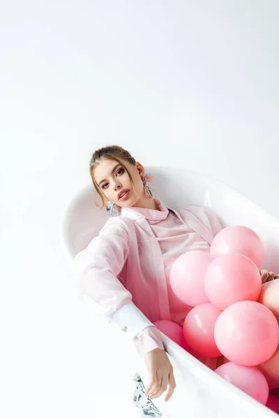 Attractive young woman lying in bathtub with pink air balloons on white — Stock Photo