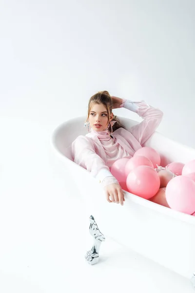 Pretty young woman lying in bathtub with pink air balloons on white — Stock Photo